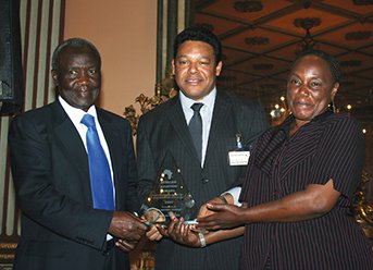 Three individuals, dressed in formal attire, stand together holding a glass award in a richly decorated setting with warm lighting.